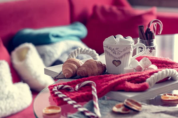 cozy winter still life, consist of, cup of coffee, marshmallow, croissants, striped lollipop on the foreground and fur shoes with  sweaters in the home interior living room