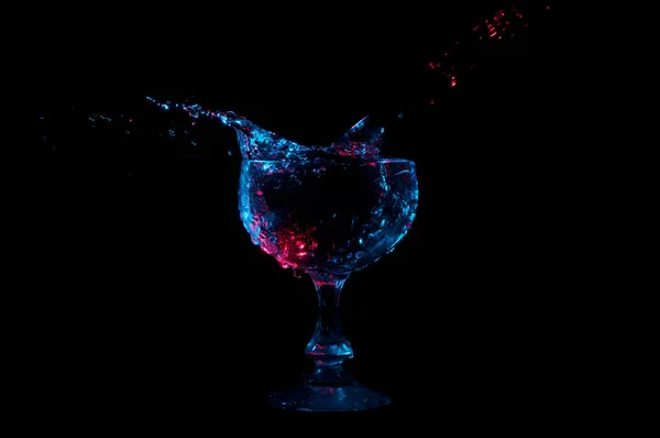 Wasser spritzt aus einem kelchförmigen Glas unter rotem und blauem Licht auf schwarzem Hintergrund — Stockfoto