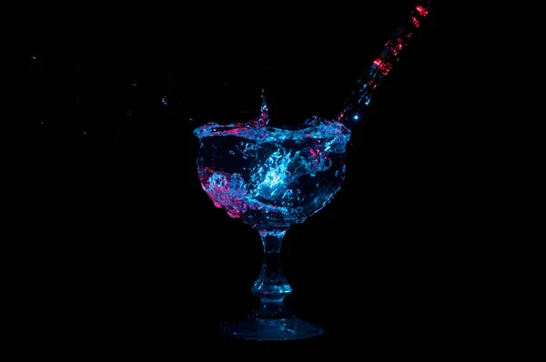 Acqua versata in un calice di vetro e spruzzata sotto le luci colorate su uno sfondo nero Foto Stock