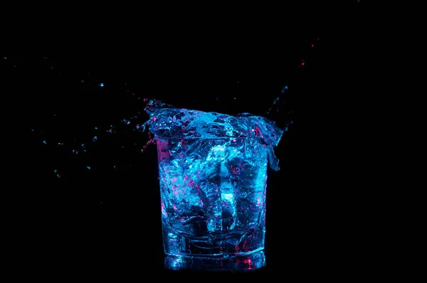 Water splashing out from a full rocks tumbler in bright blue light with red accents isolated on a black background — Stock Photo, Image