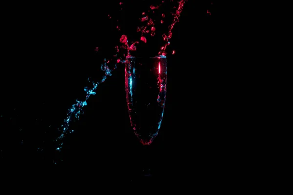 Νερό πιτσίλισμα στο μια διαγώνια γραμμή γύρω από ένα φλάουτο σαμπάνια σε κόκκινο και μπλε φωτός που απομονώνονται σε ένα μαύρο φόντο — Φωτογραφία Αρχείου