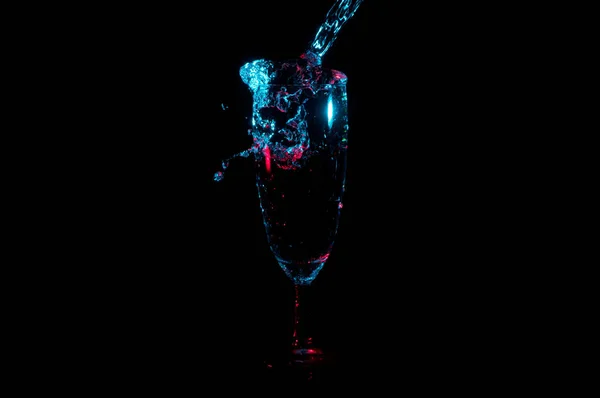 Water spilling out of tall glass under blue lights with red highlights isolated on a black background — Stock Photo, Image