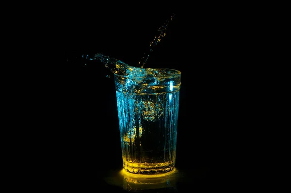 Salpicaduras de agua del vaso de beber surcado con luces azules y amarillas aisladas sobre un fondo negro — Foto de Stock