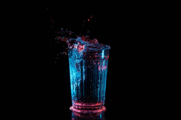 Líquido salpicando de un vidrio forrado vintage bajo luces azules y rojas aisladas sobre un fondo negro —  Fotos de Stock