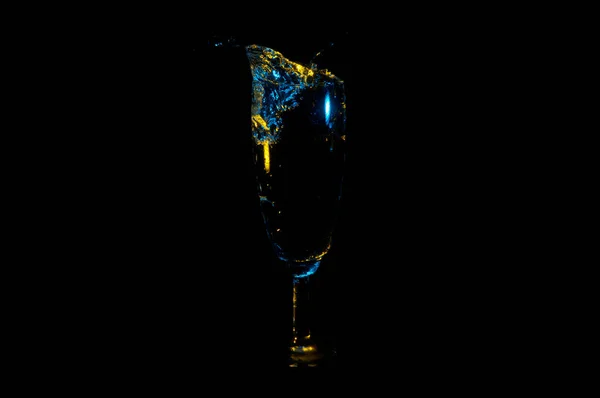 Beverage splashing out of a tall glass under blue and yellow lights isolated on a black background — Stock Photo, Image