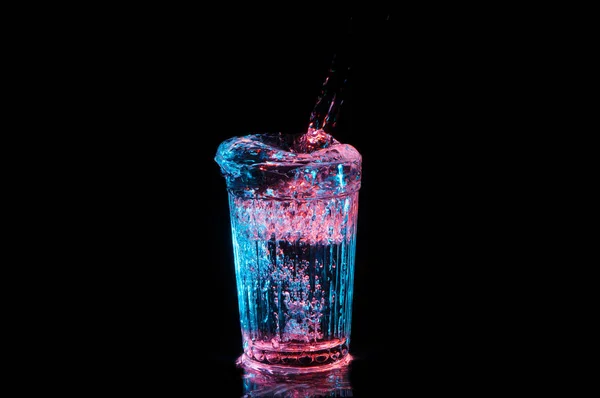 Σαφές ποτό που χύνεται σε ένα vintage ποτήρι κάτω από κόκκινο και μπλε φώτα που απομονώνονται σε ένα μαύρο φόντο — Φωτογραφία Αρχείου