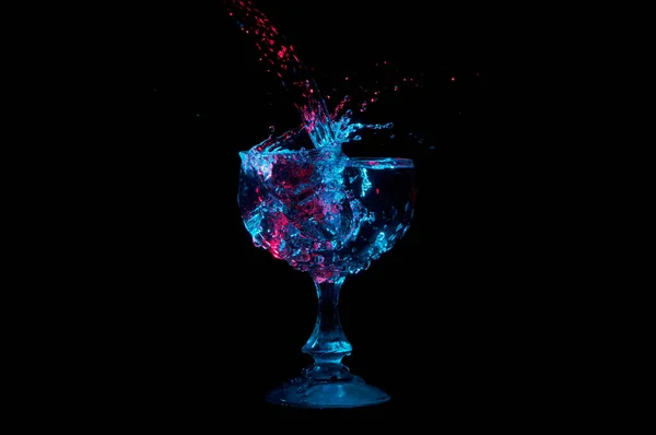 Corriente de agua salpicando en un vaso lleno bajo luces rojas y azules aisladas sobre un fondo negro — Foto de Stock