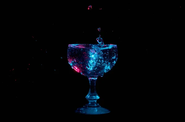 Gocce d'acqua sospese nell'aria che cadono in un calice di vetro illuminato da luci blu e rosse isolate su fondo nero — Foto Stock