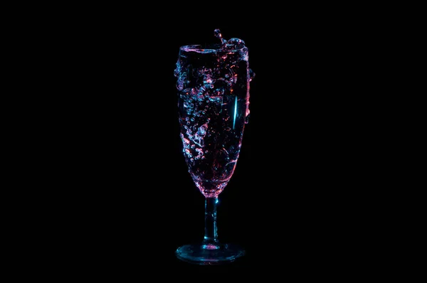 Líquido transparente que fluye sobre el borde superior de un vaso bajo luces azules y rojas aisladas sobre un fondo negro —  Fotos de Stock