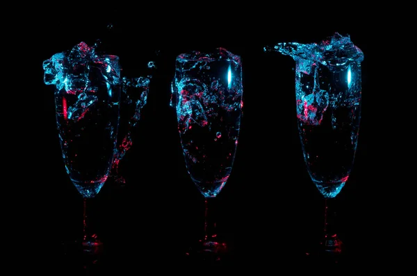 Schizzi e bolle in fila di tre bicchieri sotto le luci blu su uno sfondo nero — Foto Stock