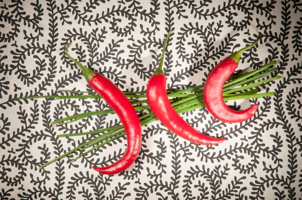 Three red chili peppers on top of a bunch of chives on a black and white patterned background
