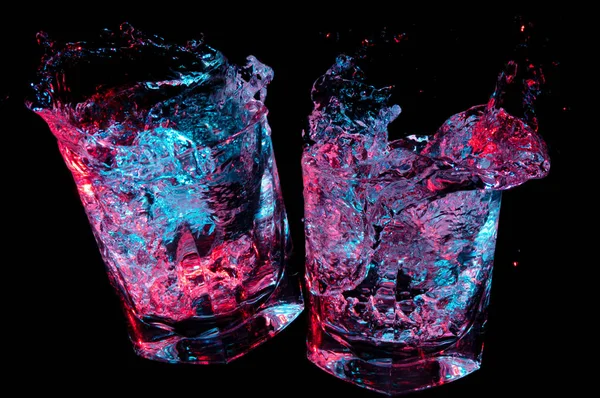 Dos vasos de rocas tintinean en tostadas mientras brilla líquido colorido salpica hacia fuera en un fondo negro — Foto de Stock