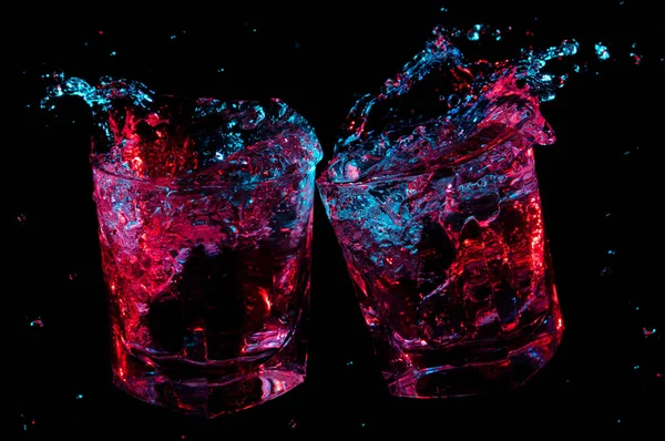 Liquido colorato incandescente schizzi da due bicchieri di rocce mentre brindano su uno sfondo nero — Foto Stock