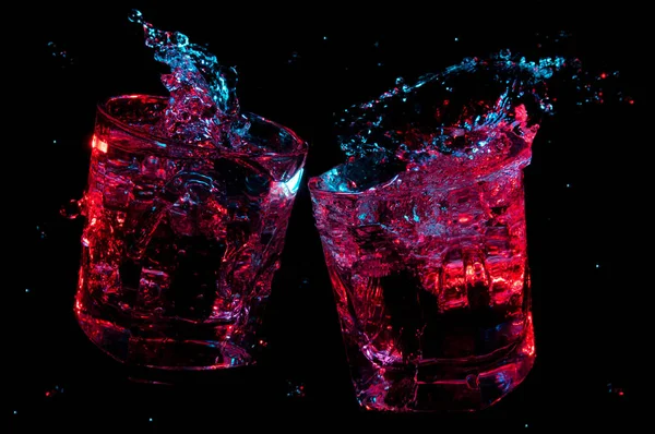 Grandes salpicaduras de líquido brillando en luces de colores en dos vasos de rocas tintineo en tostadas aisladas sobre un fondo negro —  Fotos de Stock