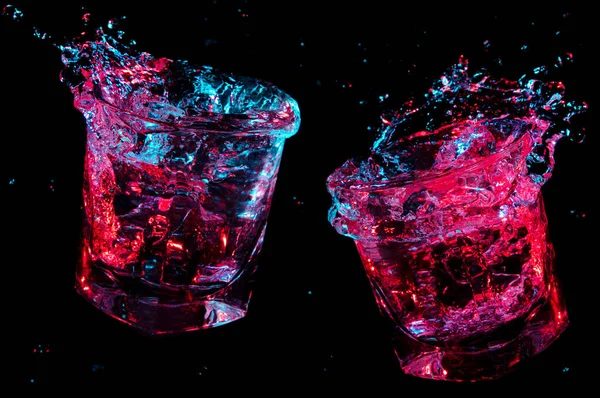 Líquido voando de dois copos de pedras cheias que acabaram de ser tostados sob luzes coloridas isoladas em um fundo preto — Fotografia de Stock