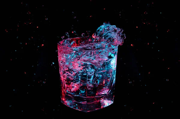 Spruzzi e goccioline di liquido in un bicchiere di rocce sotto illuminazione colorata isolato su uno sfondo nero — Foto Stock