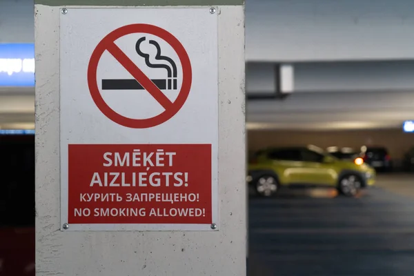 Red No smoking allower sign in three languages in an underground parking with cars visible in the background