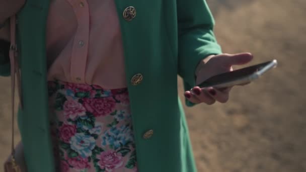 Молодая девушка достает телефон из кармана и пишет сообщение в зеленой модной куртке и красочной юбке — стоковое видео