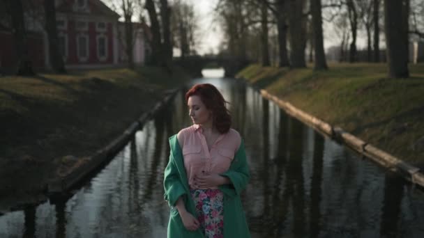 Mujer joven que se relaja cerca de un canal de la parte del palacio durante un clima cálido de primavera con chaqueta de moda verde - Pelirroja — Vídeos de Stock