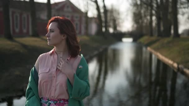 年轻女子穿着绿色时尚夹克在温暖的春天天气里在宫殿部分运河附近放松--红发 — 图库视频影像