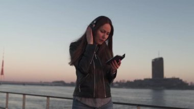 Genç bir kadın, Daugava nehrinin yakınındaki bir gün batımında deri ceket ve kot pantolon giyerek kapalı kulaklıkla müzik dinliyor.
