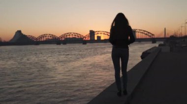 Genç bir kadın gün batımında Daugava nehri boyunca yürürken kendine güveniyor.