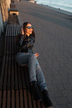 Genç bir kadın, Daugava nehrinin yakınındaki bir gün batımında deri ceket ve kot pantolon giyerek kapalı kulaklıkla müzik dinliyor.