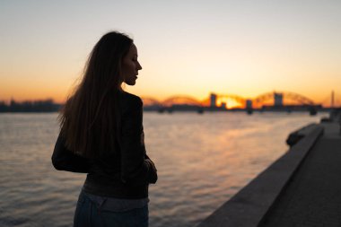 Gün batımının tadını çıkaran genç bir kadın Daugava nehri boyunca açık mavi gökyüzü manzaralı bir yürüyüş yapıyor.