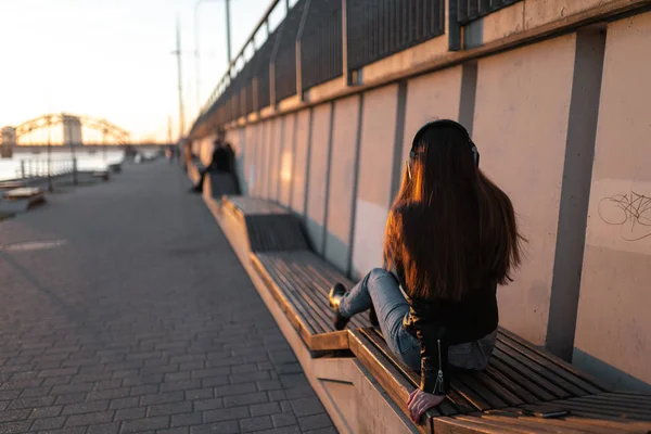 Young woman listens to music in closed headphones through her phone wearing a leather jacket and jeans at a sunset near river Daugava — 스톡 사진