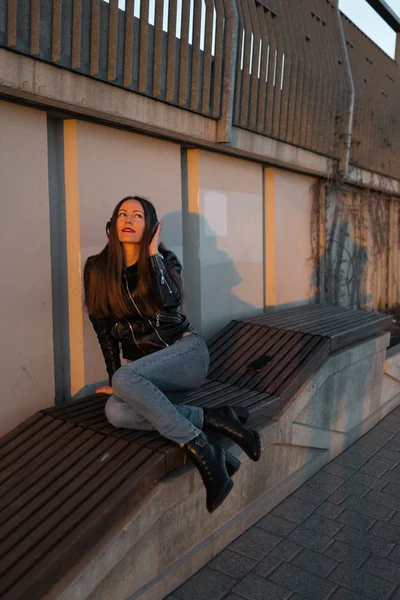 Mujer joven escucha música en auriculares cerrados a través de su teléfono con una chaqueta de cuero y jeans en una puesta de sol cerca del río Daugava — Foto de Stock