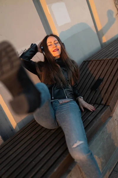 Jonge vrouw luistert naar muziek in gesloten koptelefoon via haar telefoon met een leren jas en jeans aan bij zonsondergang bij de Daugava rivier — Stockfoto