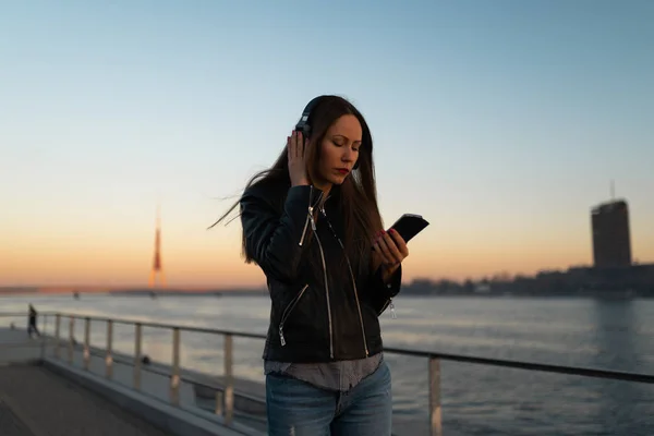 Young woman listens to music in closed headphones through her phone wearing a leather jacket and jeans at a sunset near river Daugava