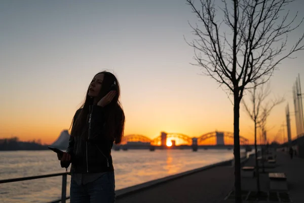 Young woman listens to music in closed headphones through her phone wearing a leather jacket and jeans at a sunset near river Daugava