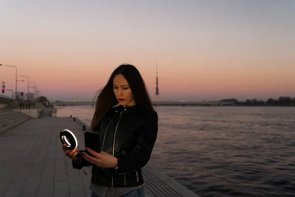 Genç bir kadın, gün batımında, Daugava nehrinin üzerinde bir manzarası olan bir ışık halkası kullanarak selfie çekiyor.