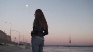Genç bir kadın gün batımında Daugava nehri boyunca yürürken kendine güveniyor.