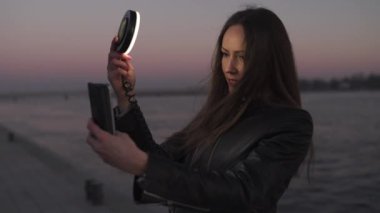 Genç bir kadın, gün batımında, Daugava nehrinin üzerinde bir manzarası olan bir ışık halkası kullanarak selfie çekiyor.