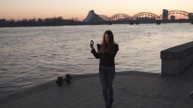 Genç bir kadın, gün batımında, Daugava nehrinin üzerinde bir manzarası olan bir ışık halkası kullanarak selfie çekiyor.