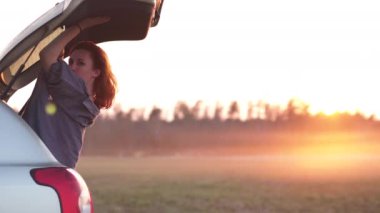 Güzel genç kadın mutlu ve Altın Saat gün batımının son dakikalarında Avrupa'da bir yolculuk sırasında bir araba bagajında dans