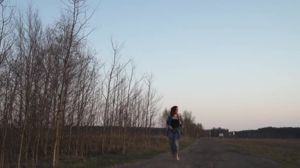 Νεαρή γυναίκα που περπατά στο πεδίο κατά τη διάρκεια ενός ηλιοβασιλέματος Golden Hour το βράδυ με καθαρό γαλάζιο ουρανό στο παρασκήνιο - Φορώντας τζιν μπουφάν και παντελόνι — Αρχείο Βίντεο
