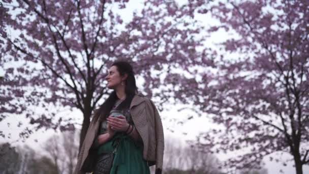 Jonge vrouw wandelen in een park met bloeiende sakura drinken van koffie uit een papieren beker dromen — Stockvideo