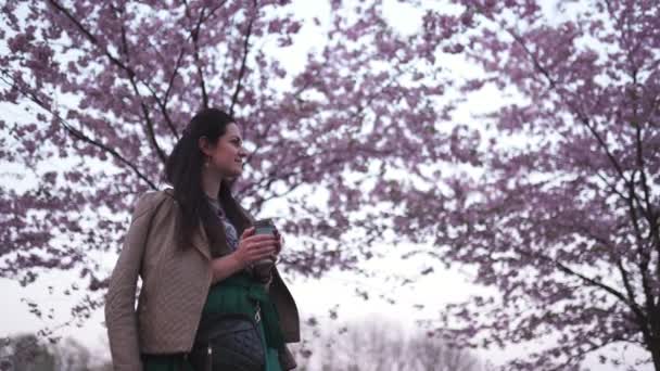 Mujer joven caminando en un parque con sakura floreciente bebiendo café de una taza de papel soñando — Vídeo de stock