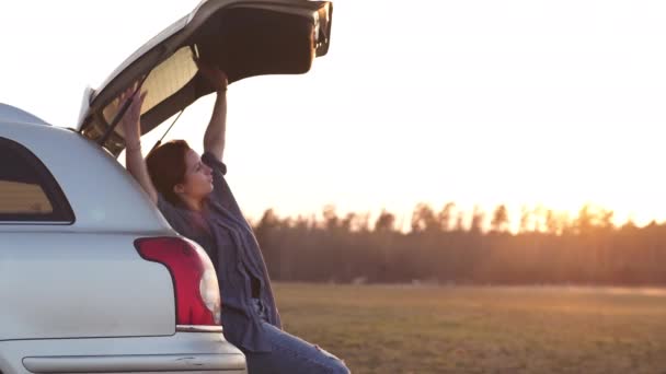Schöne junge Frau glücklich und tanzend im Kofferraum eines Autos während einer Roadtrip in Europa in den letzten Minuten der goldenen Stunde Sonnenuntergang — Stockvideo