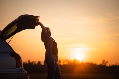 Güzel genç kadın mutlu ve Altın Saat gün batımının son dakikalarında Avrupa'da bir yolculuk sırasında bir araba bagajında dans