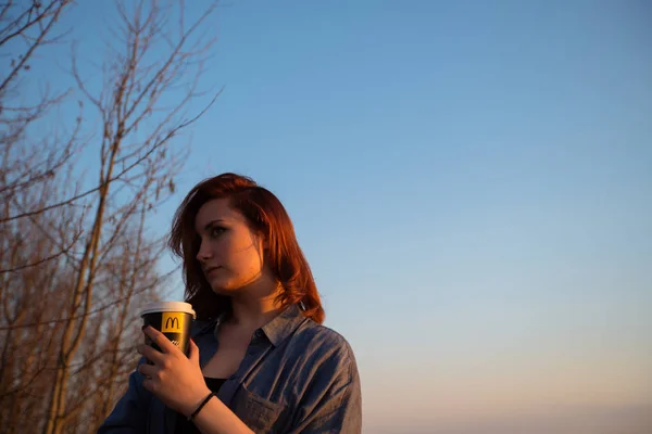 Марзно, Латвія-22 квітня 2019: молода жінка п'є каву МакДональдз на відкритому повітрі в полі під час заходу сонця — стокове фото