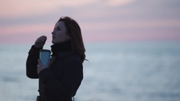 Mujer joven bebiendo tomar café al aire libre en una playa durante una puesta de sol azul frío con un claro reflejo en el agua en el Mar Báltico — Vídeos de Stock