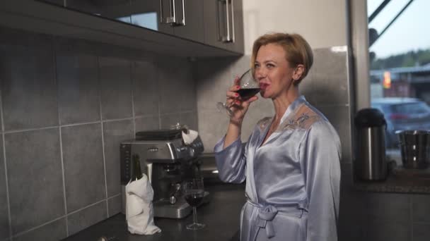 Mulher solteira bebendo vinho tinto de um copo em uma cozinha usando vestido de manhã azul - rindo e sorrindo — Vídeo de Stock