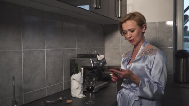 A telefonja használata közben Egyedülálló szőke nő vörös bort iszik egy pohárból a konyhában kék reggeli ruhában - Nevetés és mosolygás — Stock videók