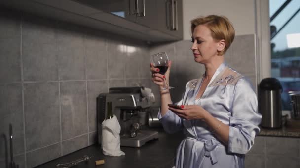 Tijdens het gebruik van haar telefoon Alleenstaande blonde vrouw drinken van rode wijn uit een glas in een keuken in blauwe ochtendjurk - Lachen en glimlachen — Stockvideo