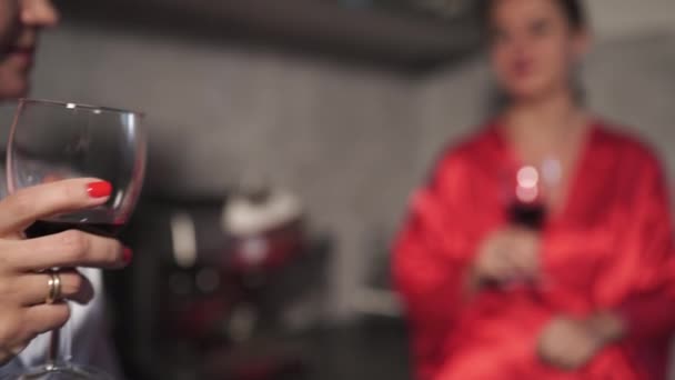 Dos mujeres charlando en la cocina y bebiendo vino tinto de cristal - Una vistiendo un vestido azul de la mañana, la otra vestido rojo - Riendo y sonriendo — Vídeo de stock