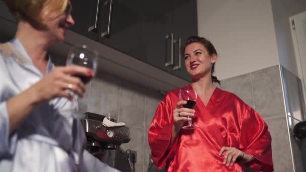 Zwei Frauen plaudern in der Küche und trinken Rotwein aus dem Glas - die eine trägt ein blaues Morgenkleid, die andere ein rotes Abendkleid - lachen und lächeln — Stockvideo